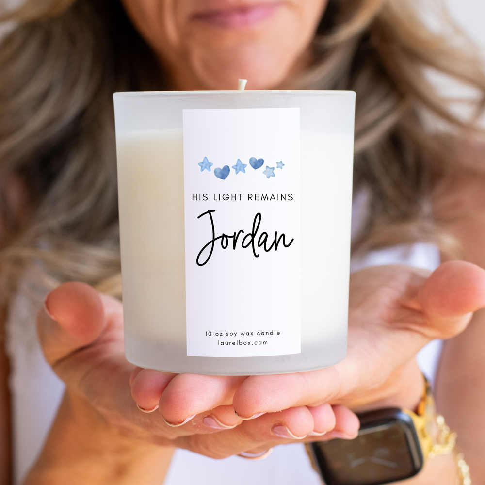 photo of hands holding a small white candle with a label that displays blue hearts and stars with the words "His Light Remains Jordan"