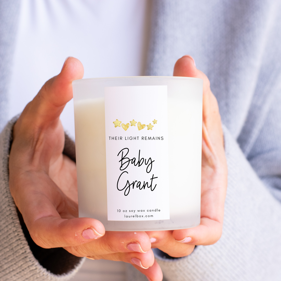photo of hands holding a small white candle with a label that display gold hearts and stars with the words "Their Light Remains Baby Grant"