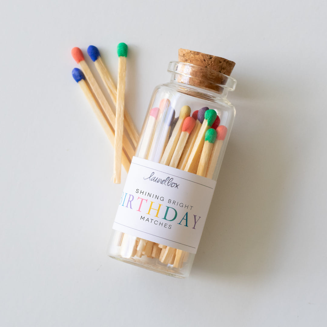 A glass container with a white label including Shining Bright Birthday Matches text filled with multi-colored matches, matches scattered on the left