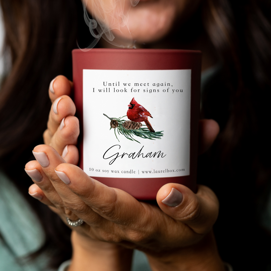 A woman holding a red-coated candle with a white sticker including the text Until we meet again, I will look for signs of you, Graham, with a cardinal image.