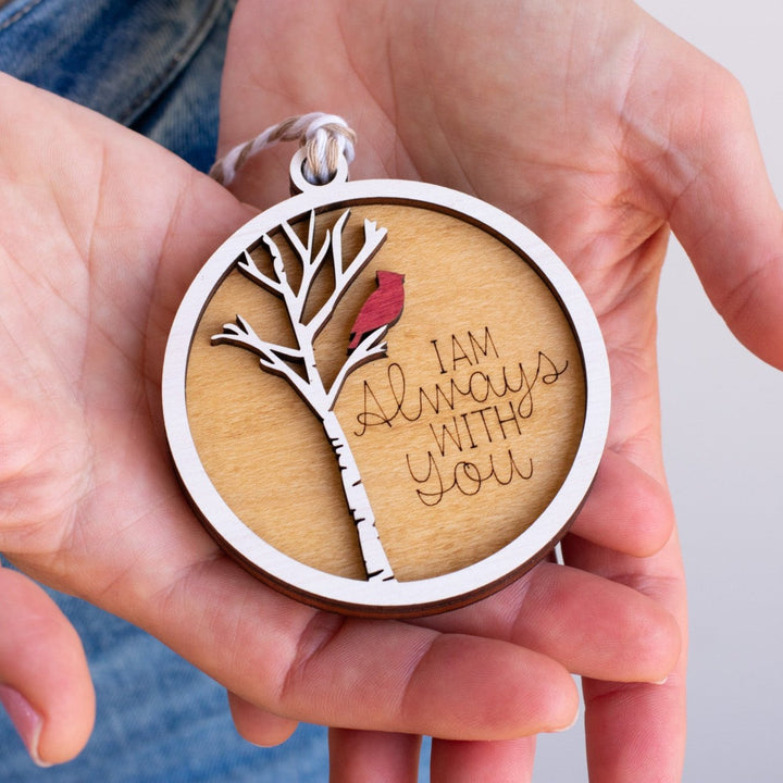 A woman is holding a maple wood ornament featuring a cardinal on a white tree,  with the text "I am always with you" engraved in both her palms.
