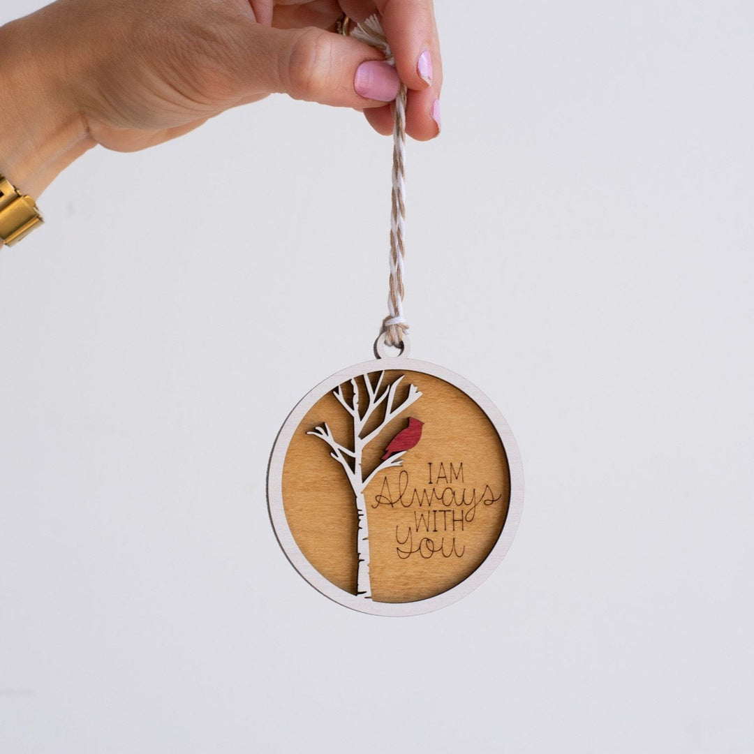 A woman holds a maple wood ornament by the thread at the top. The ornament features a cardinal on a white tree, with the text "I am always with you" engraved on it.
