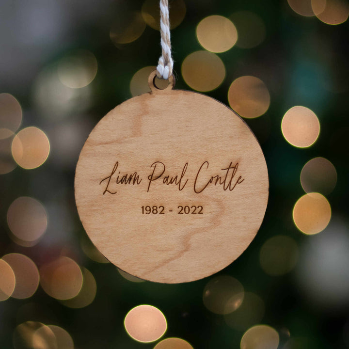 A close-up of a maple wood ornament featuring white and tan yarn at the top, with the text "Liam Paul Conette" with a blurred sparkling light background.