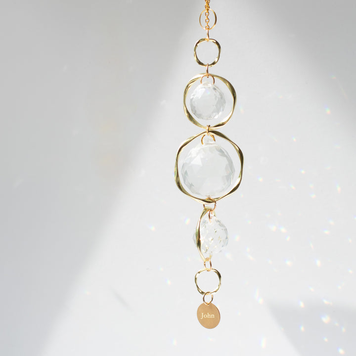 A close-up of a yellow gold sun-catcher with three crystal balls and a yellow-gold circular chain at the bottom, with the text "John" against a white background.