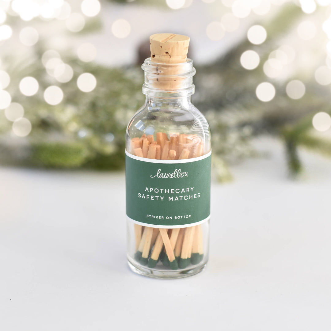 A glass container with a green label featuring the Apothecary Safety Matches text, filled with green matches, against a white background with branches and sparkling lights.