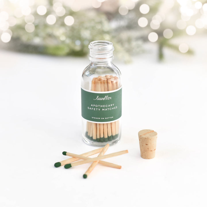 A glass container with a green label featuring the Apothecary Safety Matches text, filled with green matches, with four matches and a cork on a white background with branches and sparkling lights.