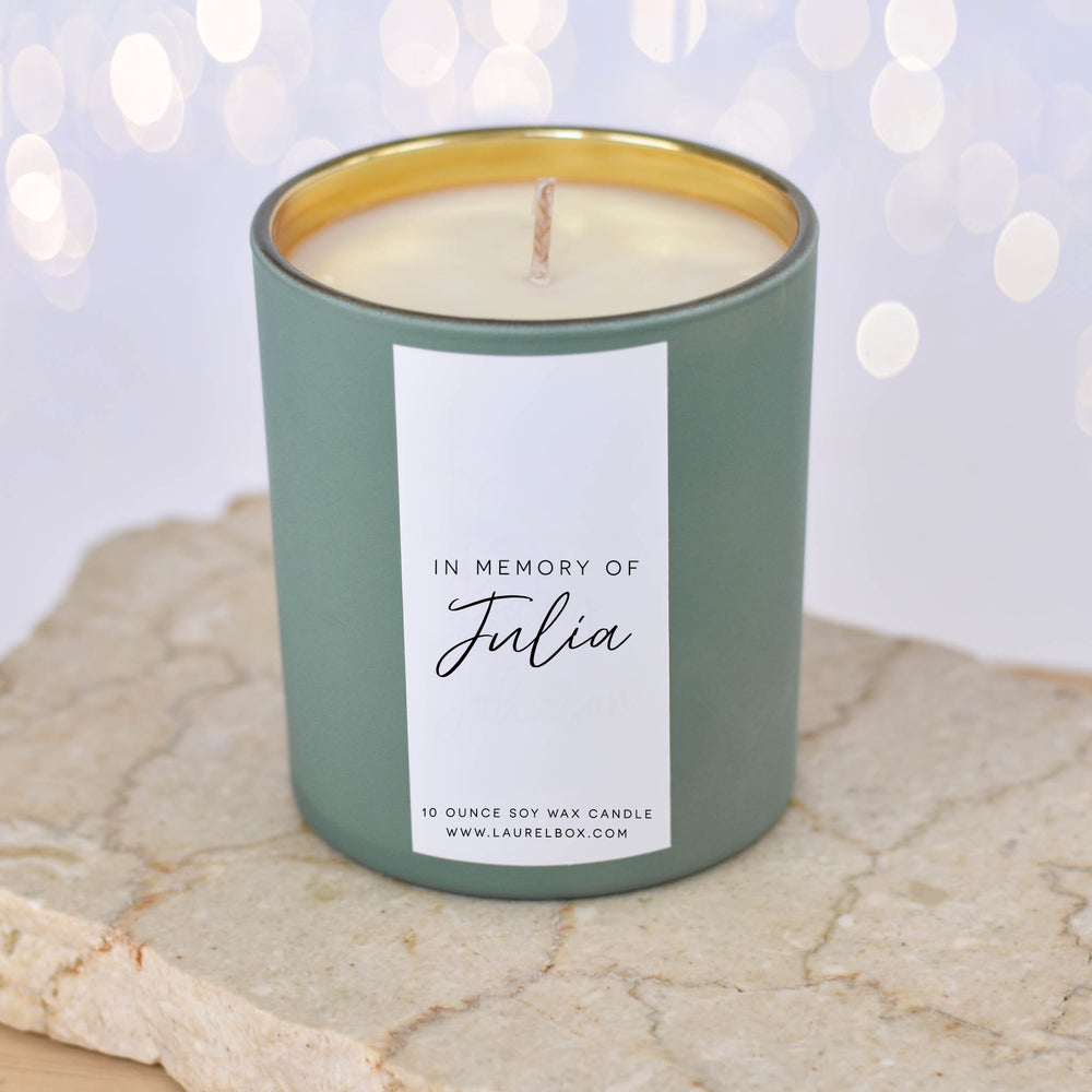 A close-up of a light green candle with the text "In Memory of Julia" on a white label sitting on a textured surface with blurred lights in the background.