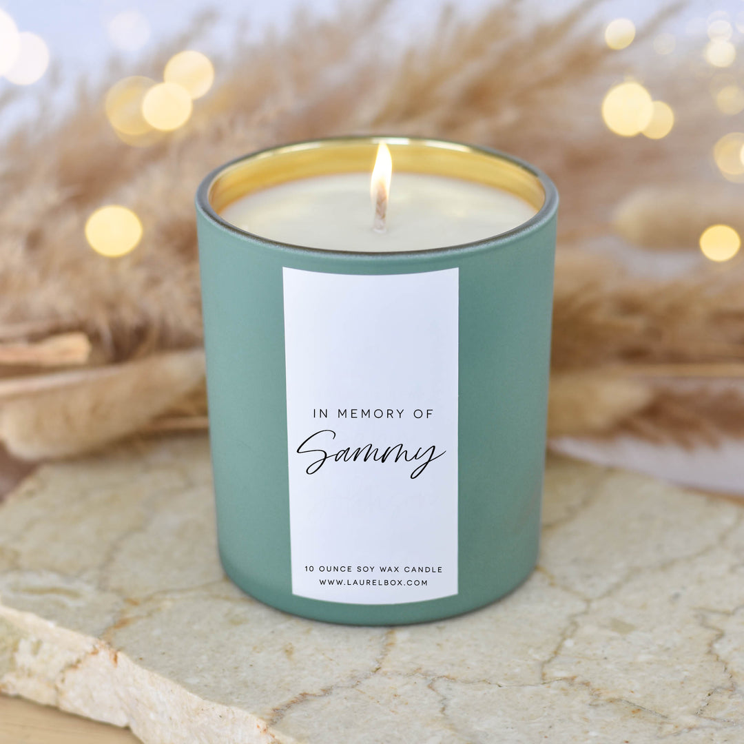 A close-up of a light green candle with the text "In Memory of Sammy" on a textured surface with blurred lights in the background.