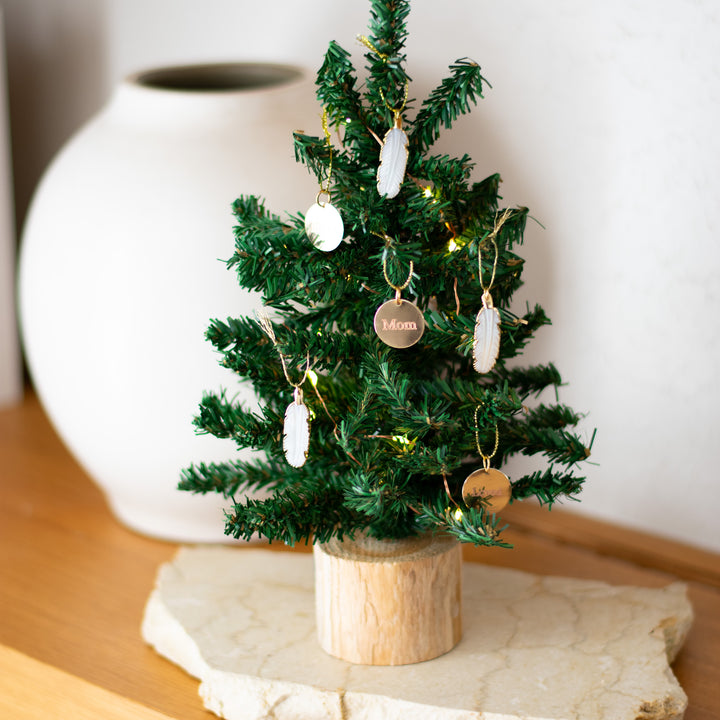 A close-up of a tabletop Christmas tree decorated with miniature feather and gold ornaments, on a wooden surface with a white vase in the background.