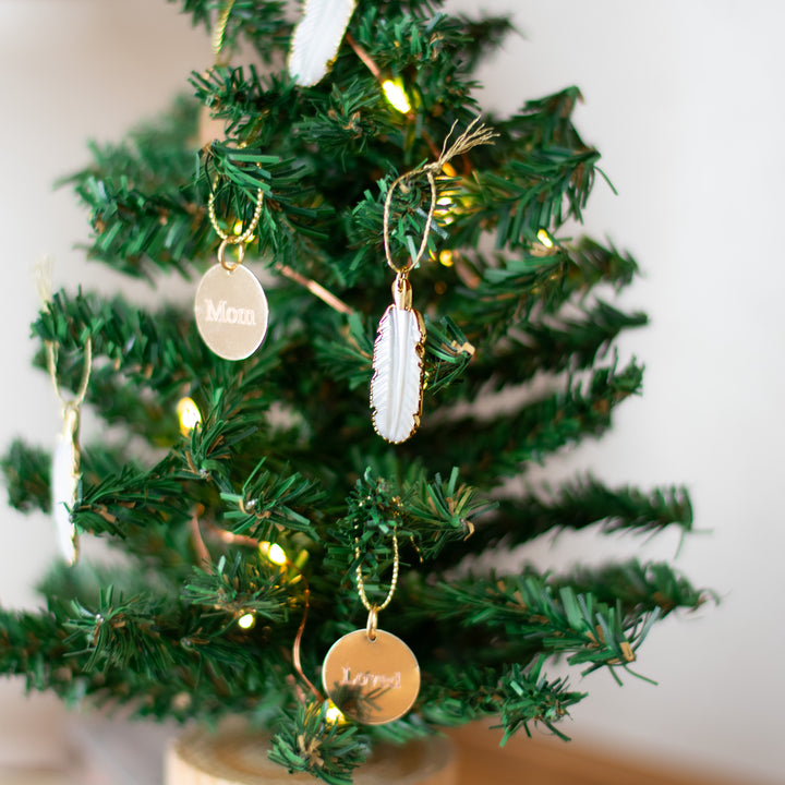 A close-up of decorative gold and white feather ornaments on a tabletop Christmas tree with blurred lights in the background.