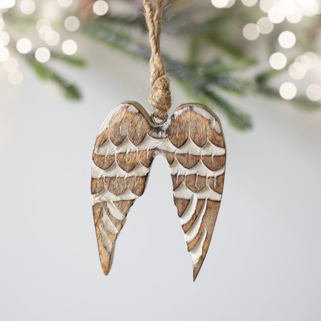 A close-up of a wooden ornament in the shape of angel wings, with natural wood color and hanging from a twine loop with Christmas tree leaves and sparkling lights blurred in the background.