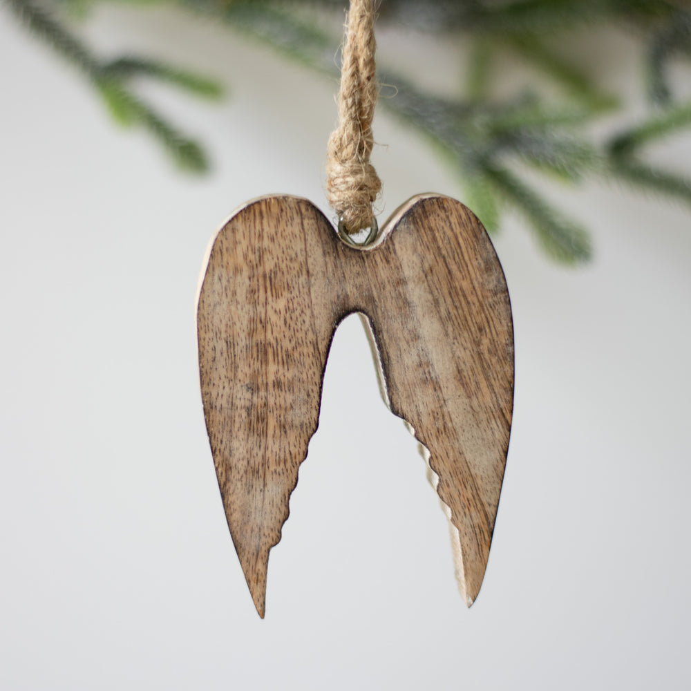 A close-up of a wooden ornament in the shape of angel wings, with natural wood color, hanging from a twine loop with Christmas tree branches blurred in the background.