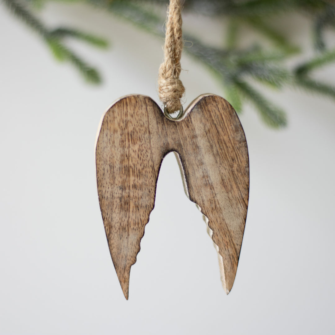 A close-up of a wooden ornament in the shape of angel wings, with natural wood color, hanging from a twine loop with Christmas tree branches blurred in the background.
