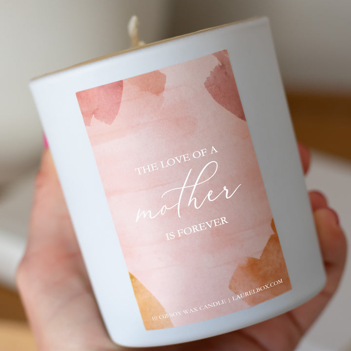 photo of a hand holding a small white candle with a pink label that displays The Love Of A Mother Is Forever in white writing