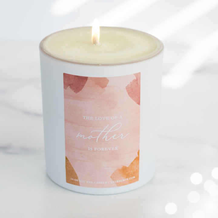 photo of a lit small white candle with a pink label that displays The Love Of A Mother Is Forever in white writing
