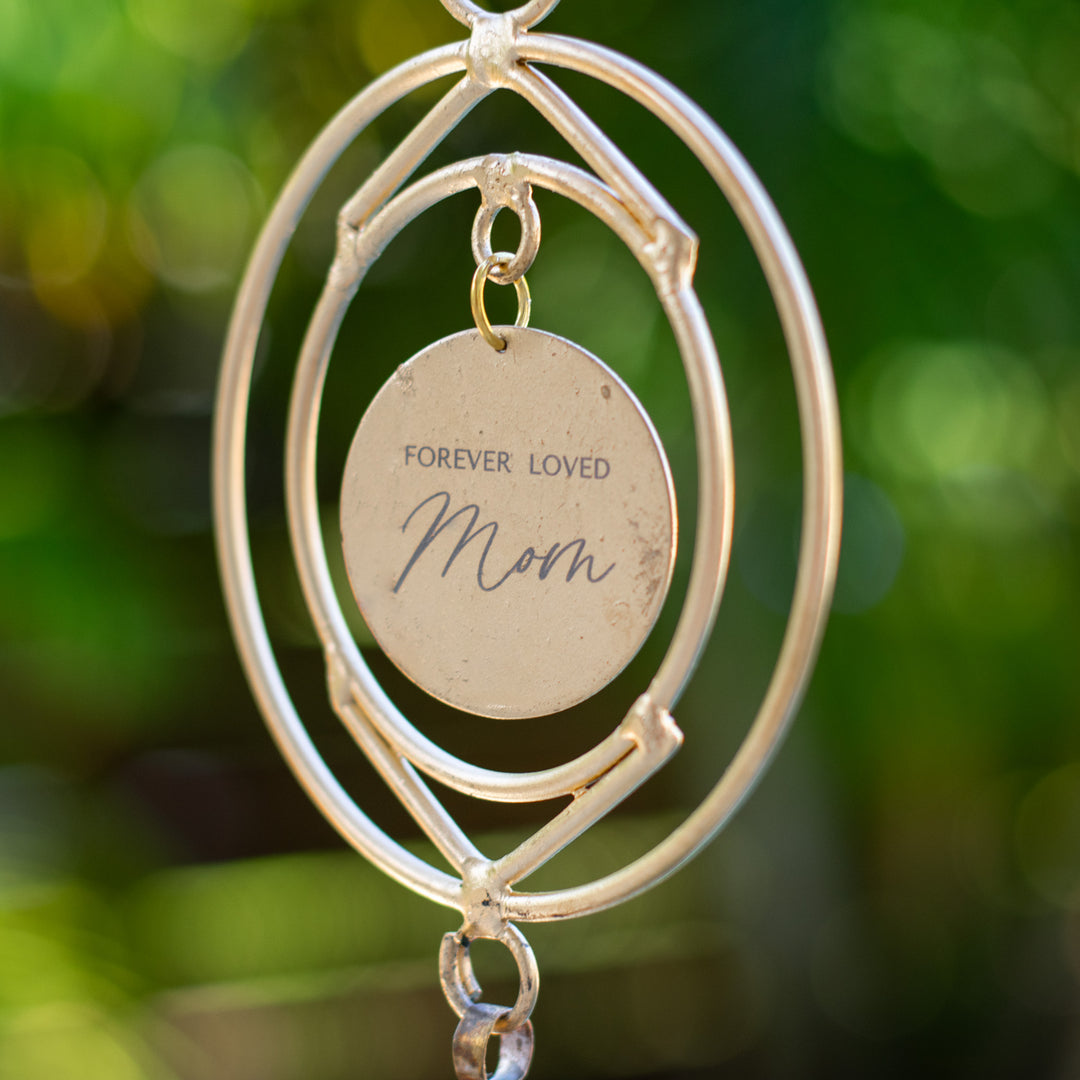 A close-up of a wind chime hanging outdoors, with a green garden background. The wind chime features two circular metal rings and a circular metal disk at the center with the engraved message: "FOREVER LOVED Mom". Below the disk, there is a small, rustic-style metal bell.