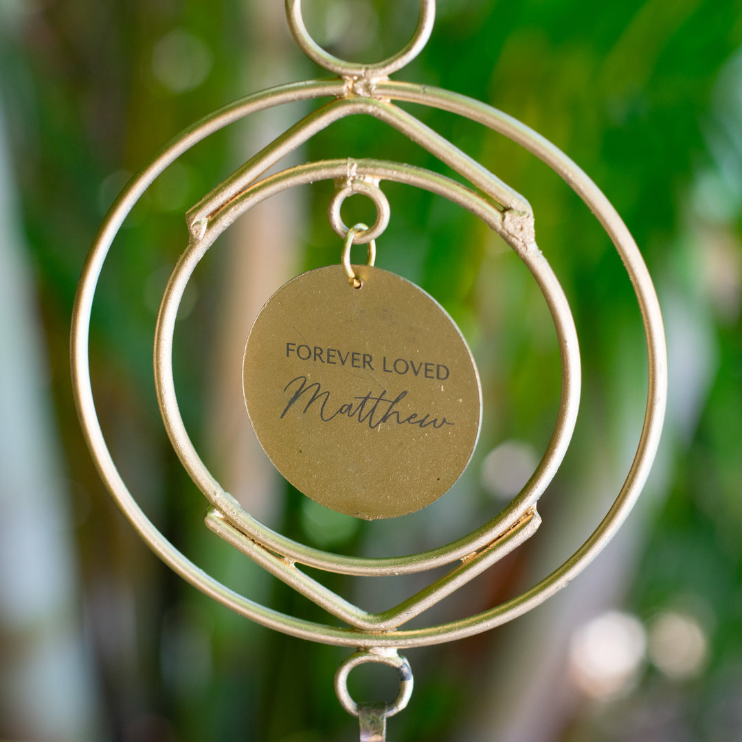 A close-up of a wind chime hanging outdoors, with a green garden background. The wind chime features two circular metal rings and a circular metal disk at the center with the engraved message: "FOREVER LOVED Matthew". Below the disk, there is a small, rustic-style metal bell.