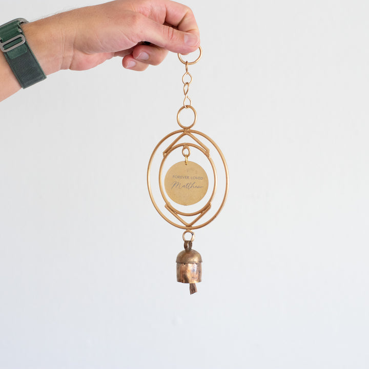 A woman is holding a wind chime against a white background. The wind chime features two circular metal rings and a circular metal disk at the center with the engraved message: "FOREVER LOVED Matthew". Below the disk, there is a small, rustic-style metal bell.