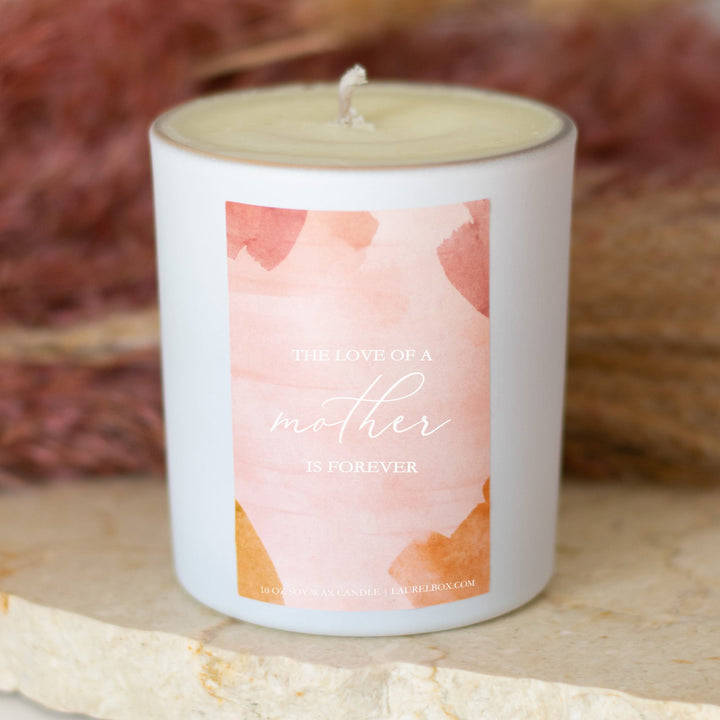 photo of a white candle with a pink label that displays The Love Of A Mother Is Forever in white writing in front of a pink background