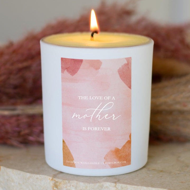 photo of a white candle with a pink label that displays The Love Of A Mother Is Forever in white writing in front of a pink and tan background