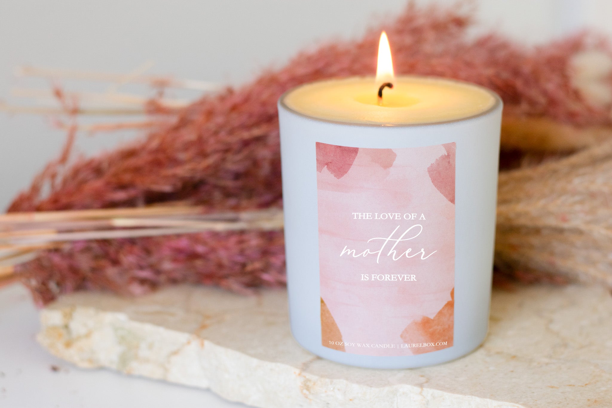 photo of a white candle with a pink label that displays The Love Of A Mother Is Forever in white writing in front of a pink and tan background