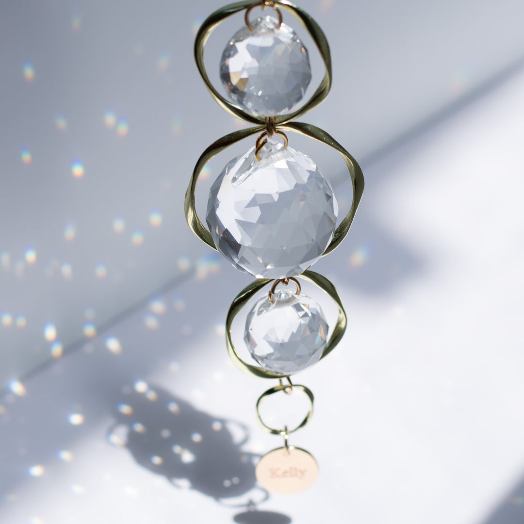 A top-view of a yellow gold sun-catcher with three crystal balls and a yellow-gold circular chain at the bottom, with the text "Kelly" against a white background.