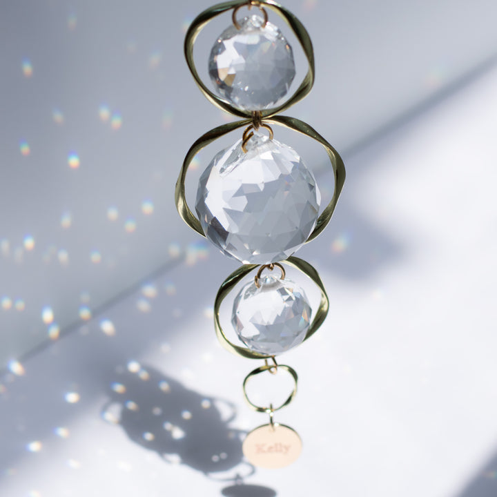 A top-view of a yellow gold sun-catcher with three crystal balls and a yellow-gold circular chain at the bottom, with the text "Kelly" against a white background.