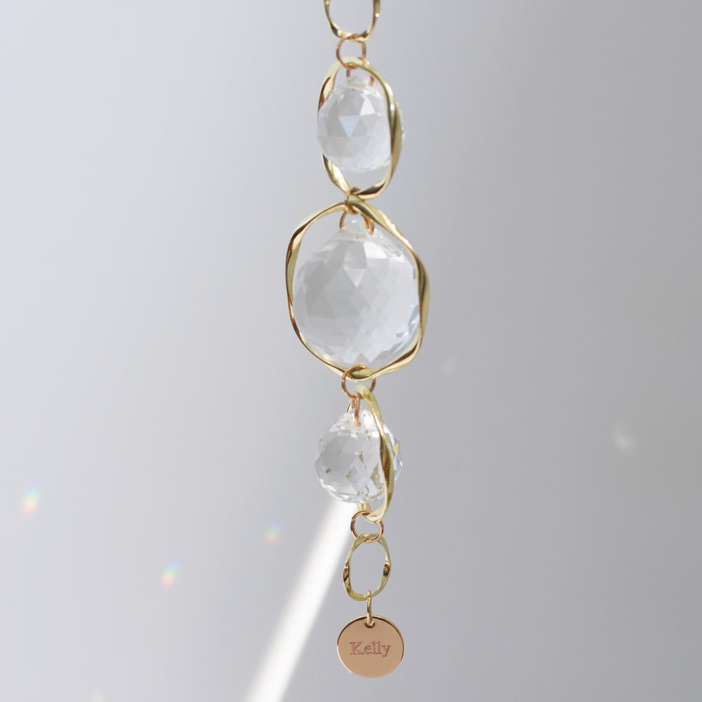 A close-up of a custom memorial sun catcher and rainbow maker, featuring an engraved charm at the bottom with the text 'Kelly' against a gray background.