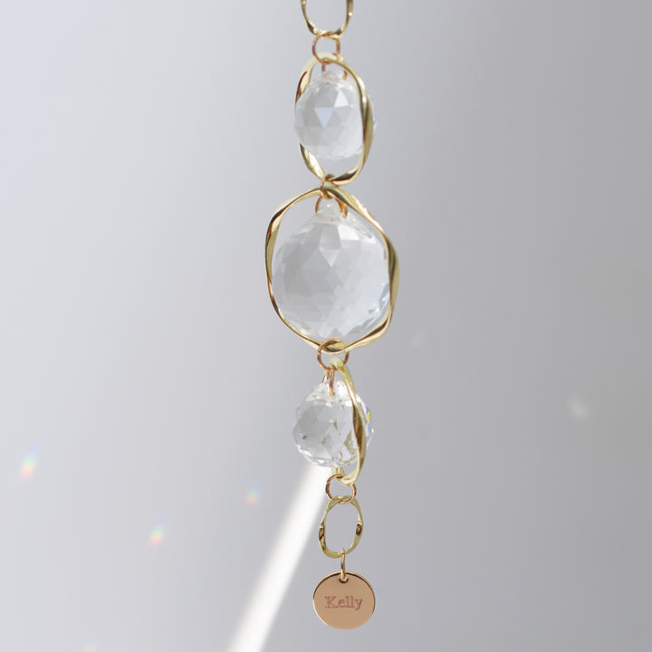 A close-up of a custom memorial sun catcher and rainbow maker, featuring an engraved charm at the bottom with the text 'Kelly' against a gray background.