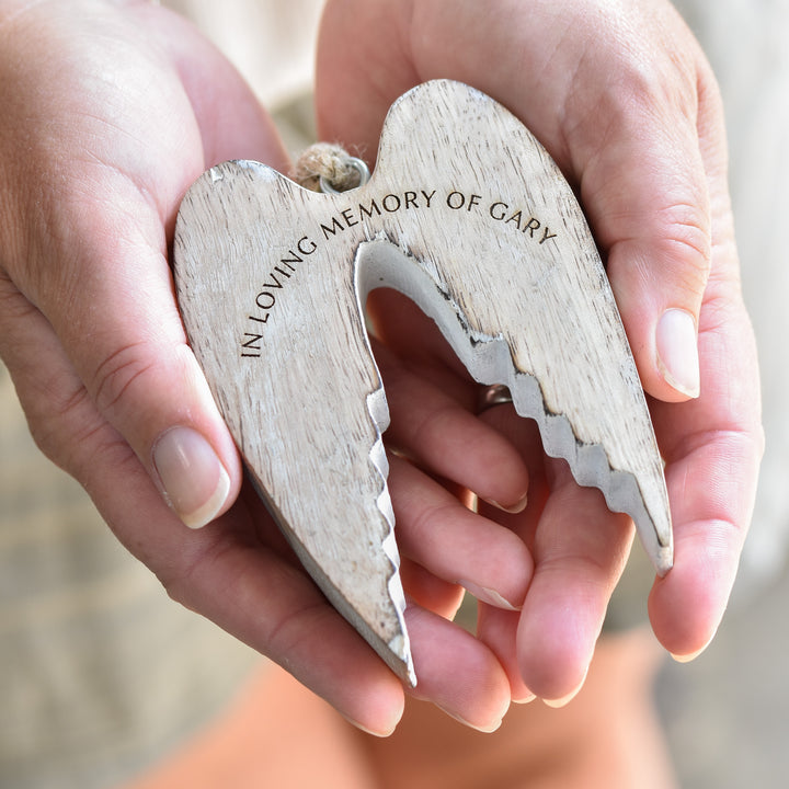 A close-up of a woman holding a white wooden ornament in the shape of angel wings, with natural wood color, with the text "In Loving Memory of Gary" and hanging with twine thread in both of her hands.