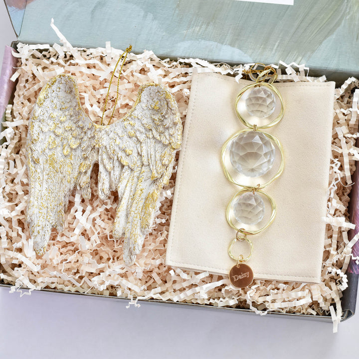 A close-up of a laurelbox gift set with an angel wing ornament with gold accents, with a gold string at the top on the left, and a gold sun-catcher with three crystal balls and a yellow-gold circular chain at the bottom, with the text "Daisy" in a box filled with tan shredded paper.