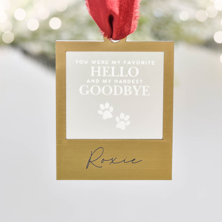 A close-up of a gold square ornament with red silk ribbon at the top, with text 'You were my favorite hello and my hardest goodbye' and paw prints, and the text "Roxie" hanging against a blurred background.