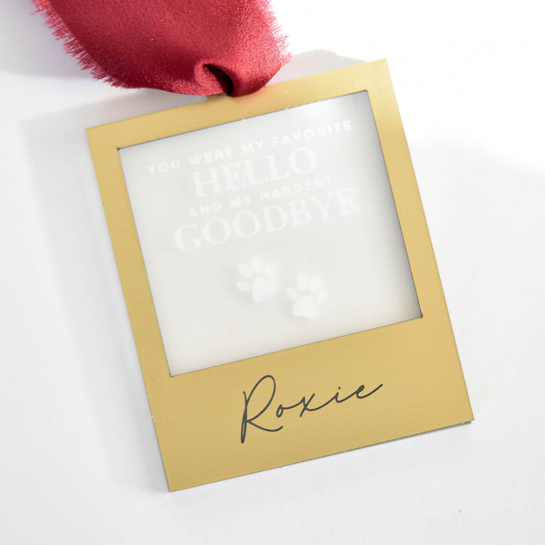 A close-up of a gold square ornament with red silk ribbon at the top, with text 'You were my favorite hello and my hardest goodbye' and paw prints, and the text "Roxie" against a white background.