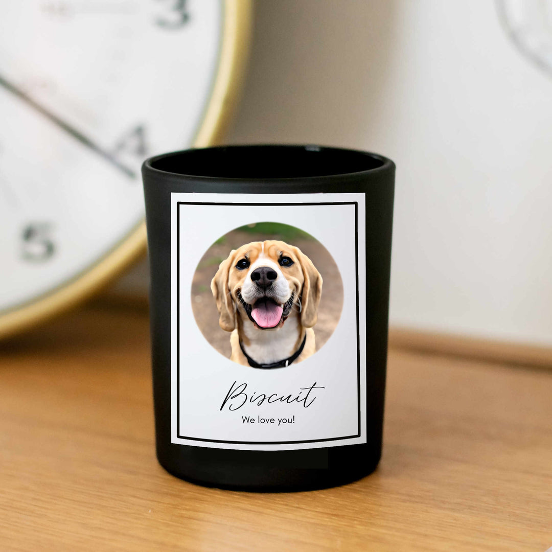 A black-coated candle with a white sticker including the text Biscuit we love you and a picture of a dog. Sitting on a wooden table with a clock behind it