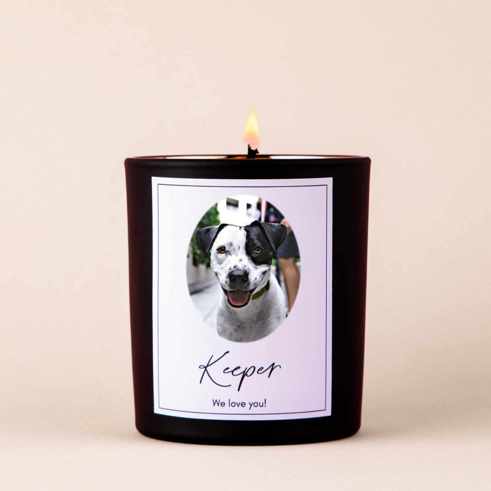 A lit black-coated candle with a white sticker including the text Keeper we love you! and a picture of a dog.