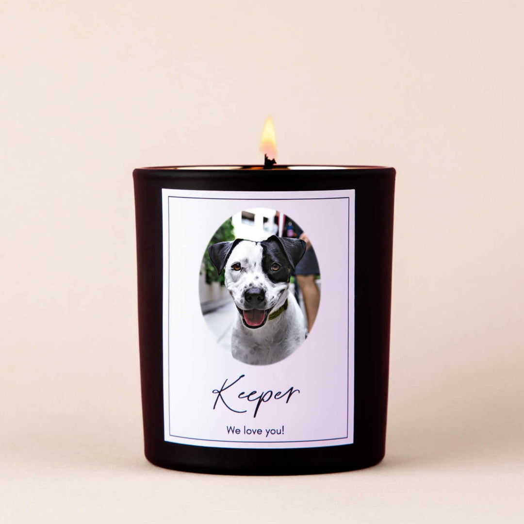 A lit black-coated candle with a white sticker including the text Keeper we love you! and a picture of a dog.