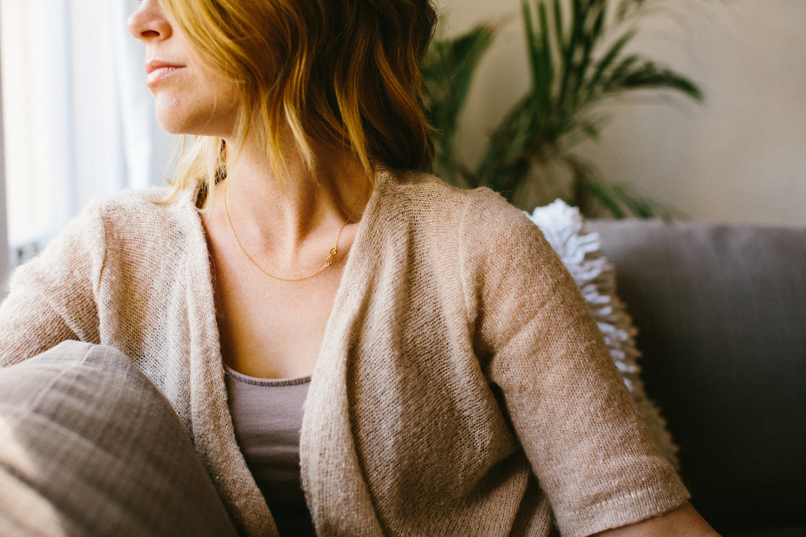 photo of a woman with blonde hair sitting on grey couch wearing a tan elbow length cardigan with a brown tank top underneath and a gold necklace with an infinity symbol on it