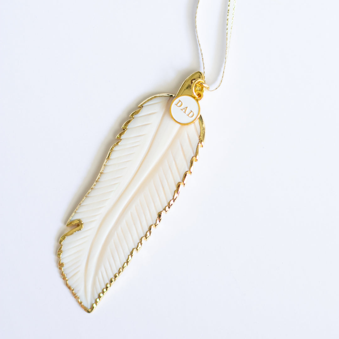 A close-up of a white mother-of-pearl feather ornament with gold edges and details and a "DAD" white and gold enamel charm at the top of the ornament.
