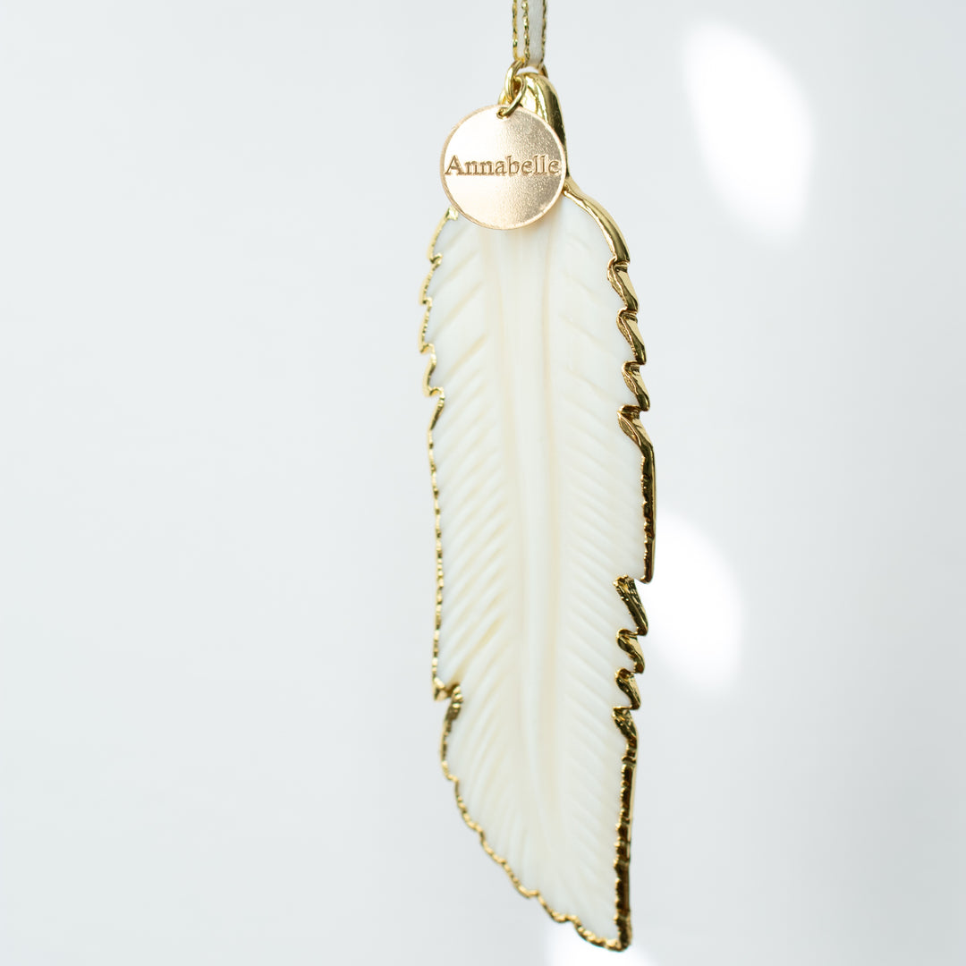A close-up of a white mother-of-pearl feather ornament with gold edges and details and an "Annabelle" white and gold enamel charm at the top of the ornament. Highlighting the details and charm.