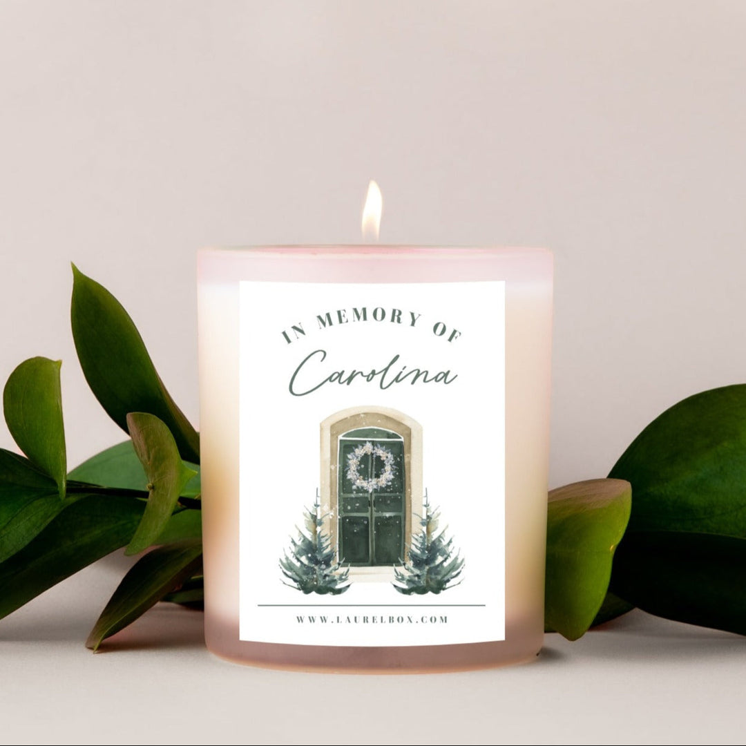 A pink coated candle with a white sticker with the text "In Memory of Carolina" and two Christmas trees and a door below the text against a white background