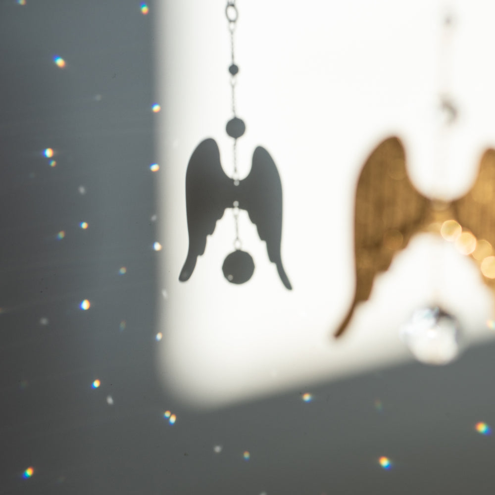 photo of the shadow of a gold chain with gold angel wings with clear crystals above and below the angel wings on a white wall with small rainbows all around it
