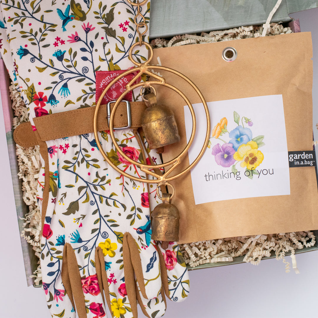 An open gift box shows a small rustic wind chime with two bells and golden metal circles, garden gloves, and a brown bag with a Thinking of you sticker with flowers and garden in a bag text on the right.