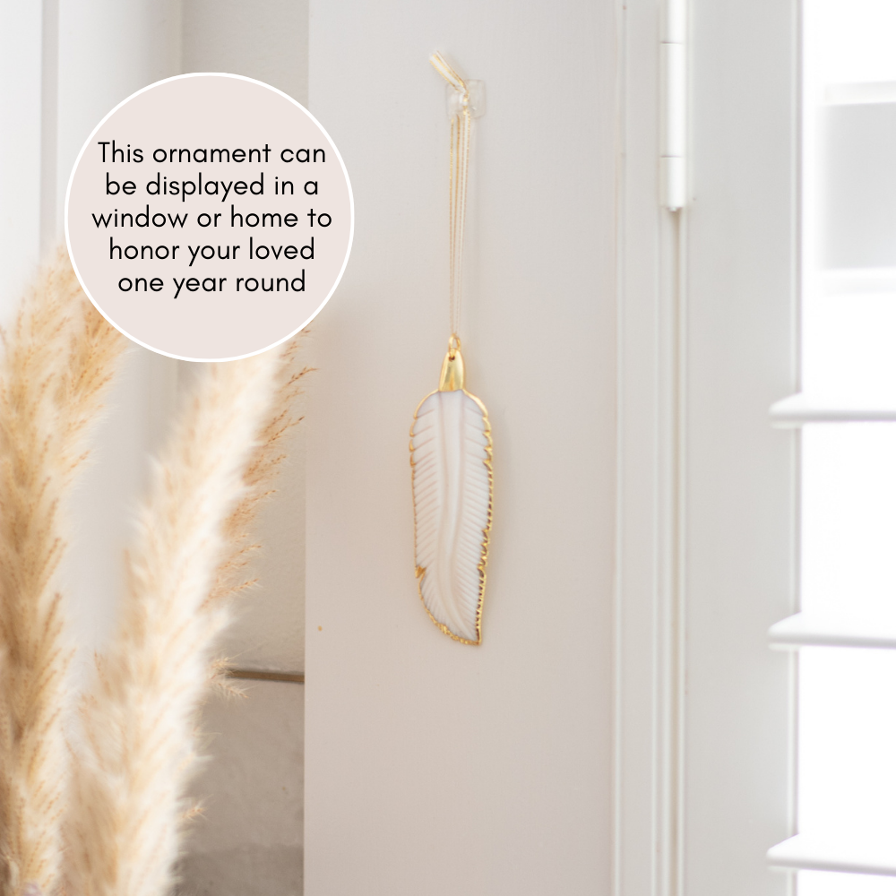 A white mother-of-pearl feather ornament hanging on a wall. Pink circle text with "this ornament can be displayed in a window or home to honor your loved one year-round" on the top left corner.