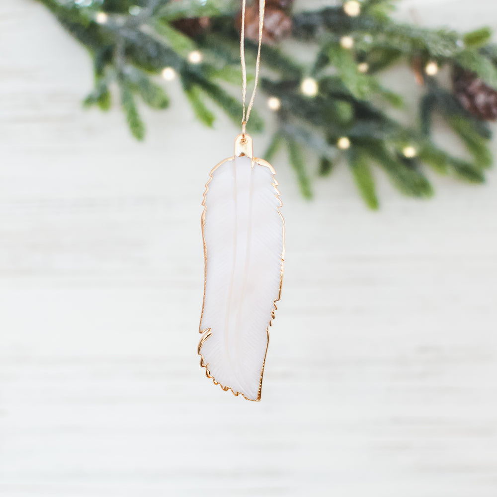A close-up of a white feather ornament with a gold outline against a white background with Christmas tree branches.