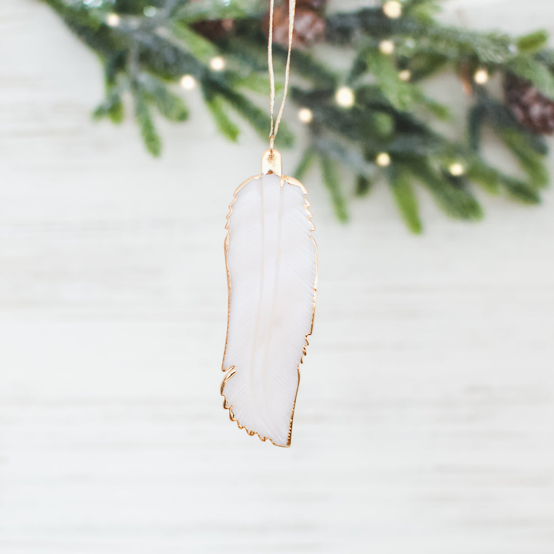 A close-up of a white feather ornament with a gold outline against a white background with Christmas tree branches.