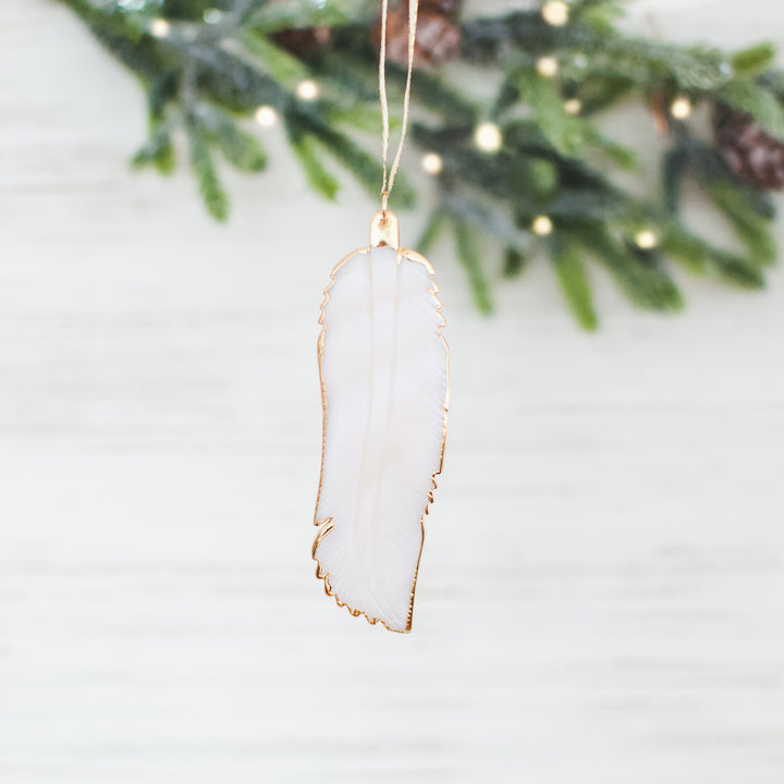 A close-up of a white feather ornament with a gold outline against a white background with Christmas tree branches.