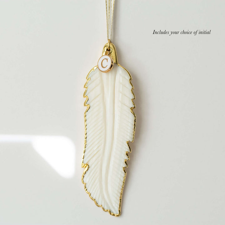A close-up of a white mother-of-pearl feather ornament with gold edges and details and a "C" initial white and gold enamel charm at the top of the ornament. With the text "includes your choice of initial" at the top right corner.