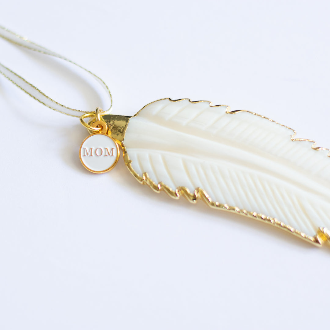 A close-up of a white mother-of-pearl feather ornament with gold edges and details, and a "MOM" white and gold enamel charm at the top of the ornament.