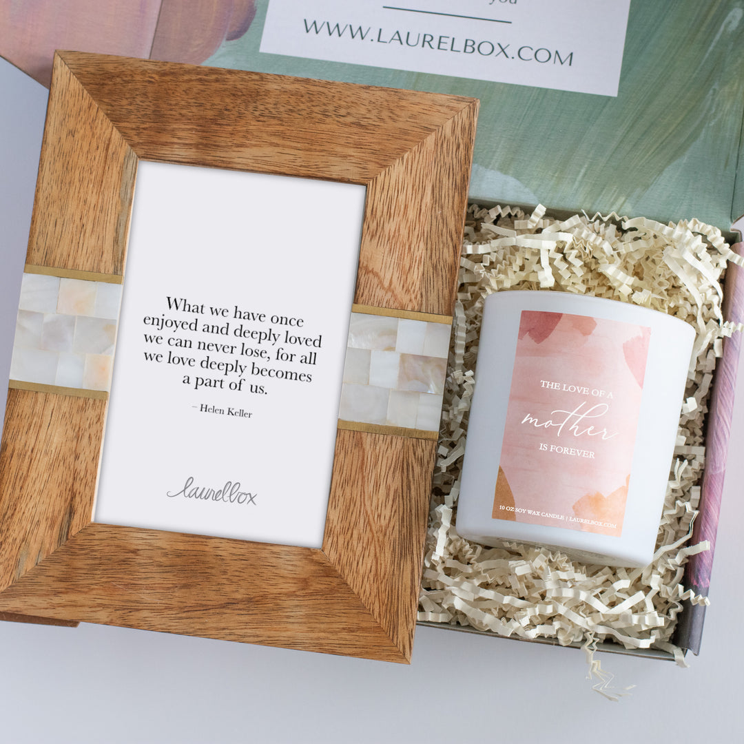 photo a of a gift box containing a wooden photo frame with a white mosaic pattern on either side of the frame and a white piece of paper displaying "What we have once enjoyed and deeply loved we can never lose, for all we love deeply becomes a part of us - Helen Keller" sitting next to a small white candle with a pink label display " The Love Of a Mother Is Forever"