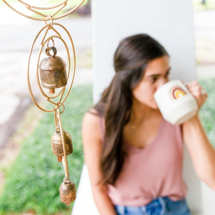 Close-up of a rustic wind chime of recycled tin and iron coated in copper with four bells and a woman blurred in the background drinking from a mug with a rainbow